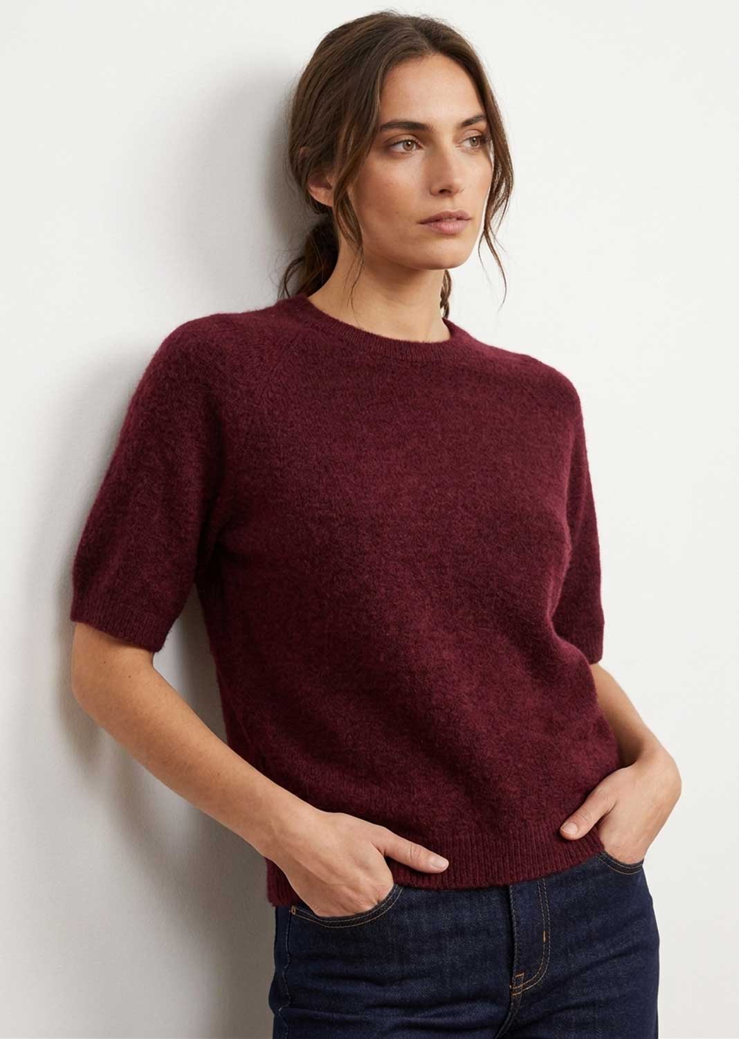 A woman wearing a short-sleeve crewneck knit sweater in deep burgundy, paired with dark blue jeans, leaning against a plain white wall.