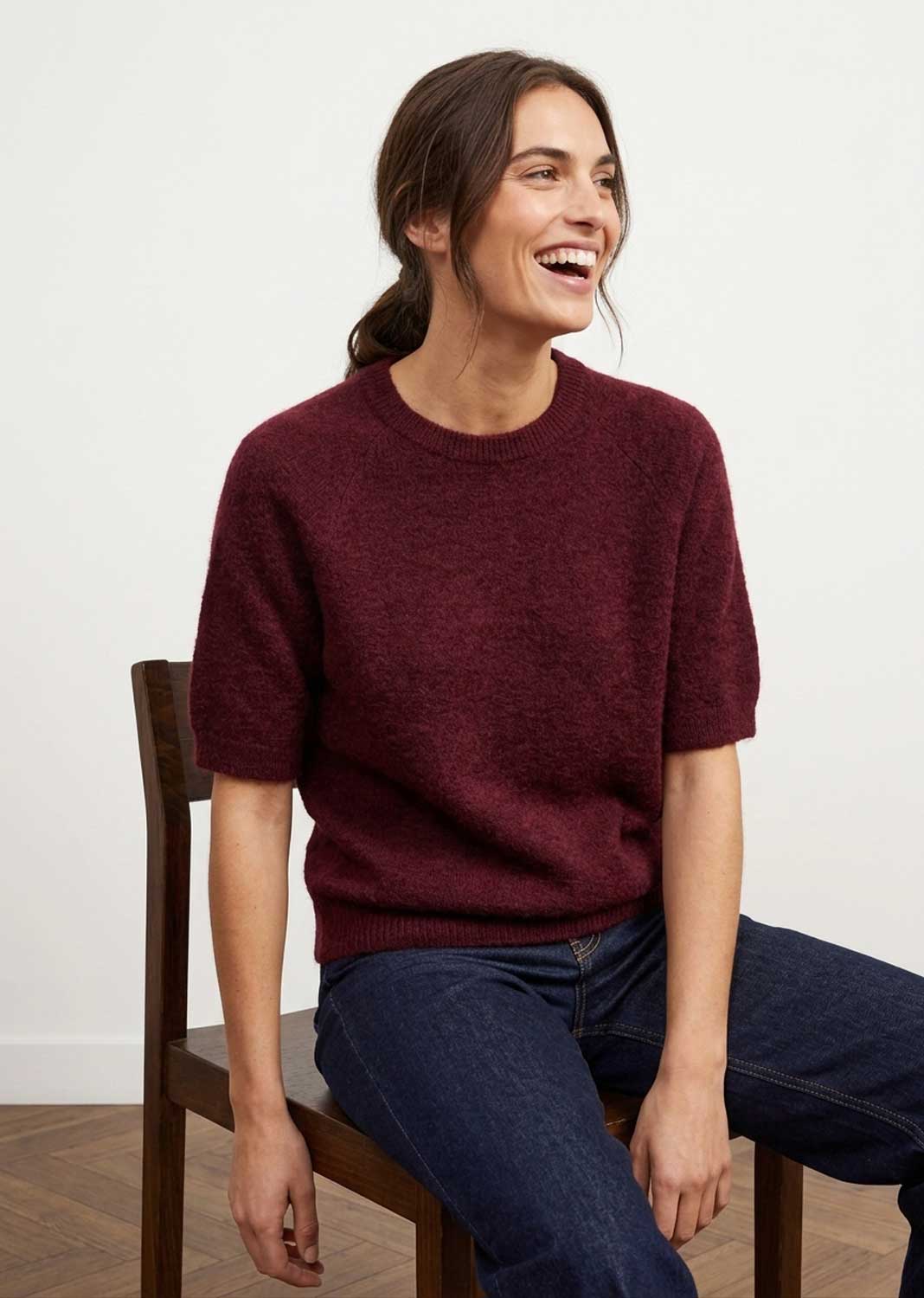 A woman sitting on a wooden chair wearing a short-sleeve crewneck knit sweater in burgundy and dark blue jeans, set against a plain white wall.
