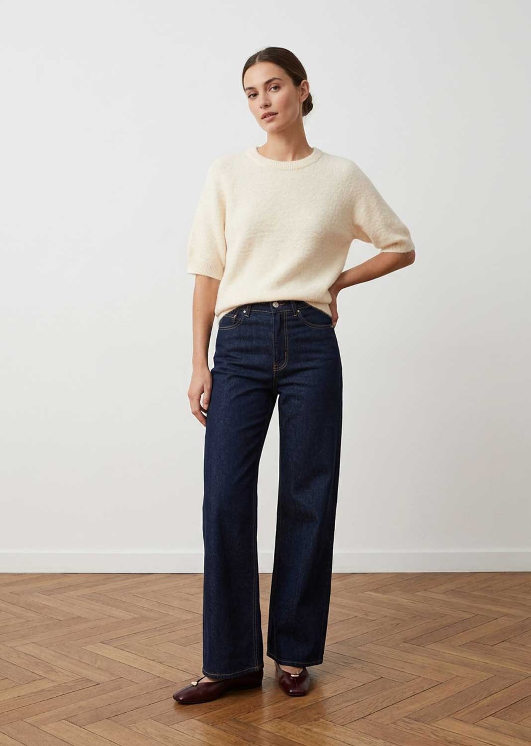 A woman stands in a minimalist room with a light wall and wood parquet floor, wearing a cream short-sleeve knit sweater, dark blue wide-leg jeans, and dark brown loafers.