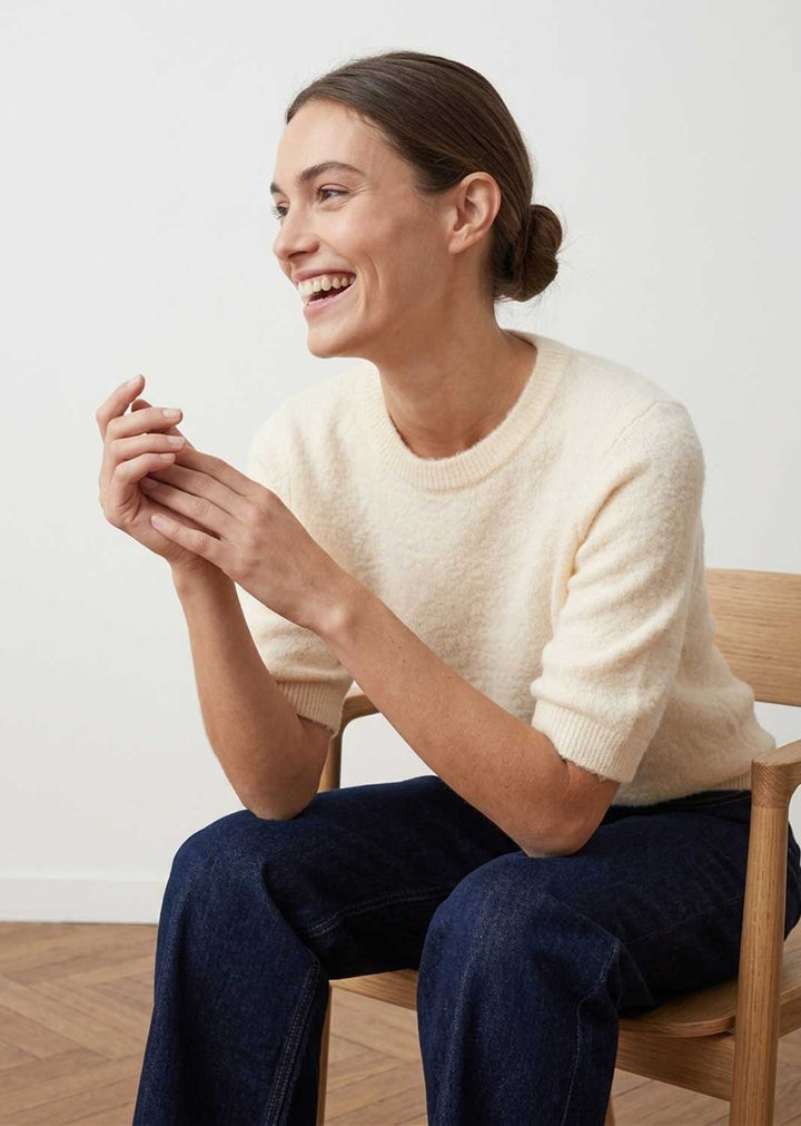 A woman sitting on a wooden chair, wearing a cream-colored short-sleeve knit sweater and dark blue jeans, smiling and looking to the side against a plain white wall.