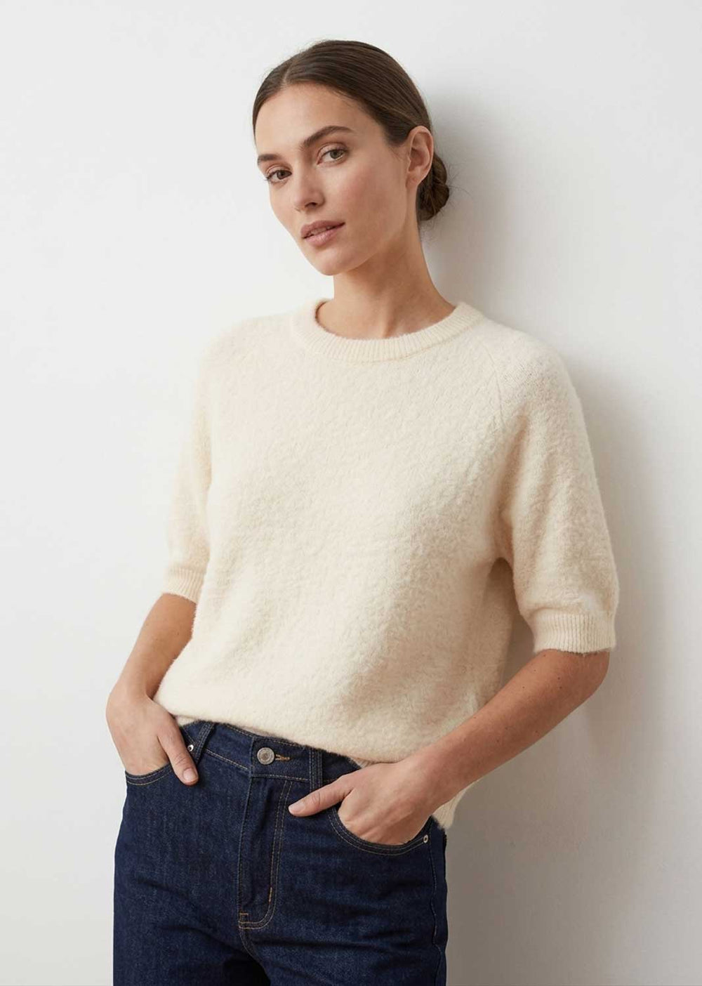 A woman wearing a cream-colored, short-sleeved fuzzy knit sweater and dark blue jeans, standing against a plain white wall.
