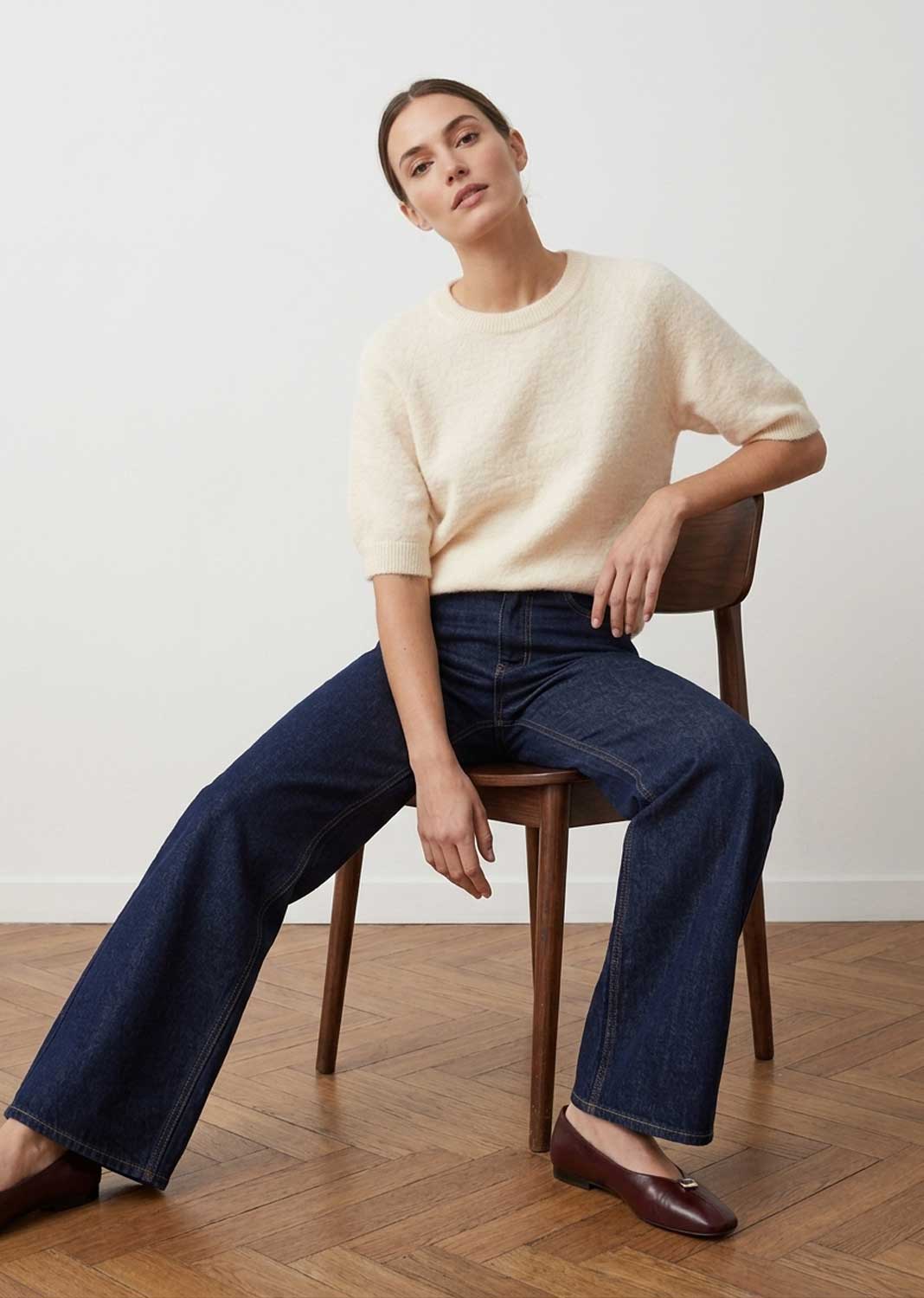 A woman sits on a wooden chair in a studio setting, wearing a cream short-sleeve knit sweater, dark blue wide-leg jeans, and dark brown flats against a white wall and wood floor.