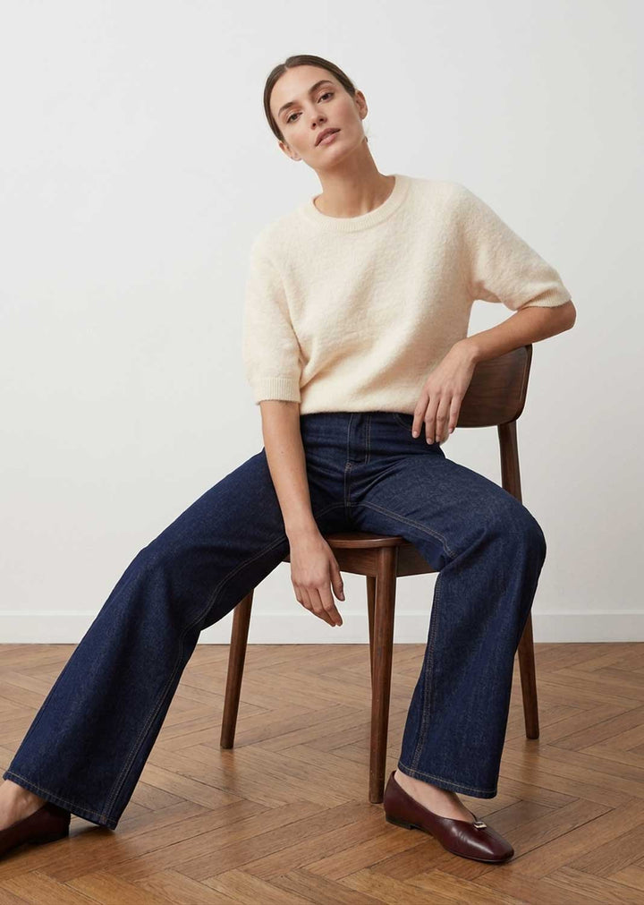A woman sits on a wooden chair in a studio setting, wearing a cream short-sleeve knit sweater, dark blue wide-leg jeans, and dark brown flats against a white wall and wood floor.