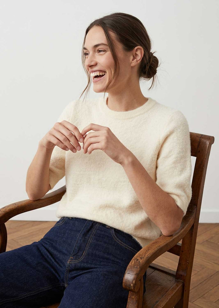 A woman smiling while sitting in a wooden armchair, wearing a cream-colored short-sleeve knit sweater and dark blue jeans against a plain light background.