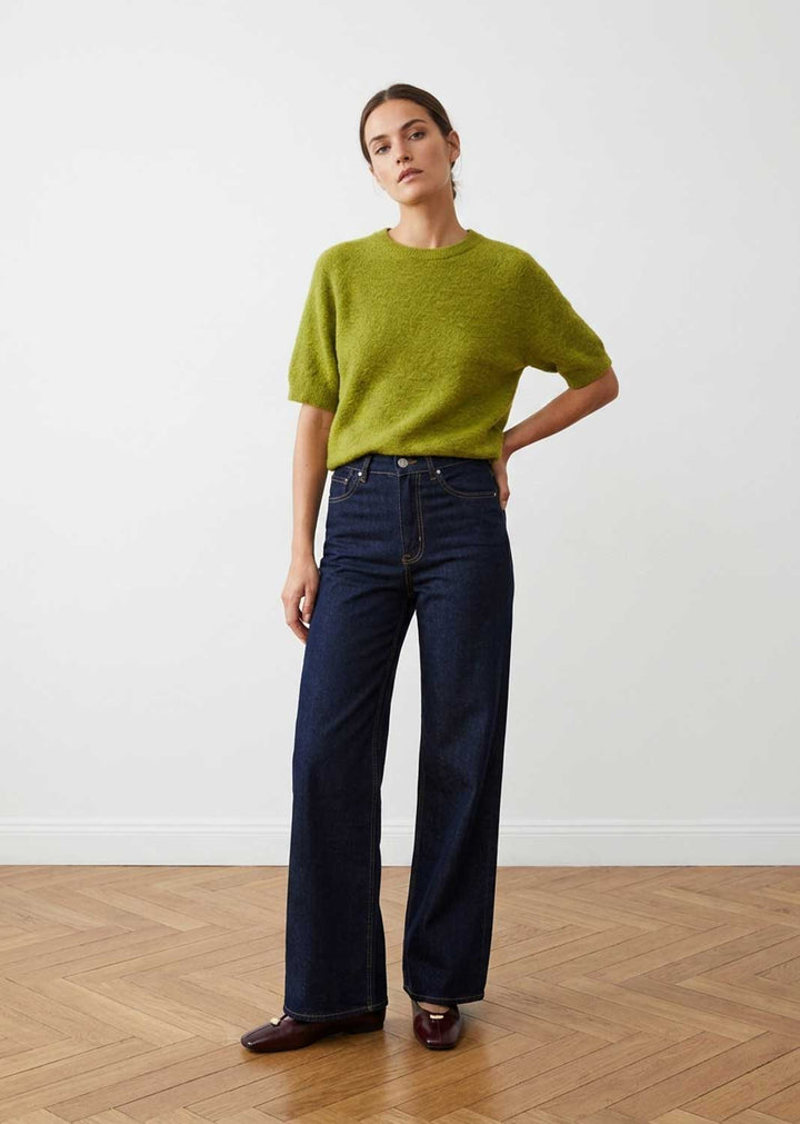 A woman modeling a short-sleeve olive green knit sweater and dark blue wide-leg jeans, standing on a light wood herringbone floor against a white wall.