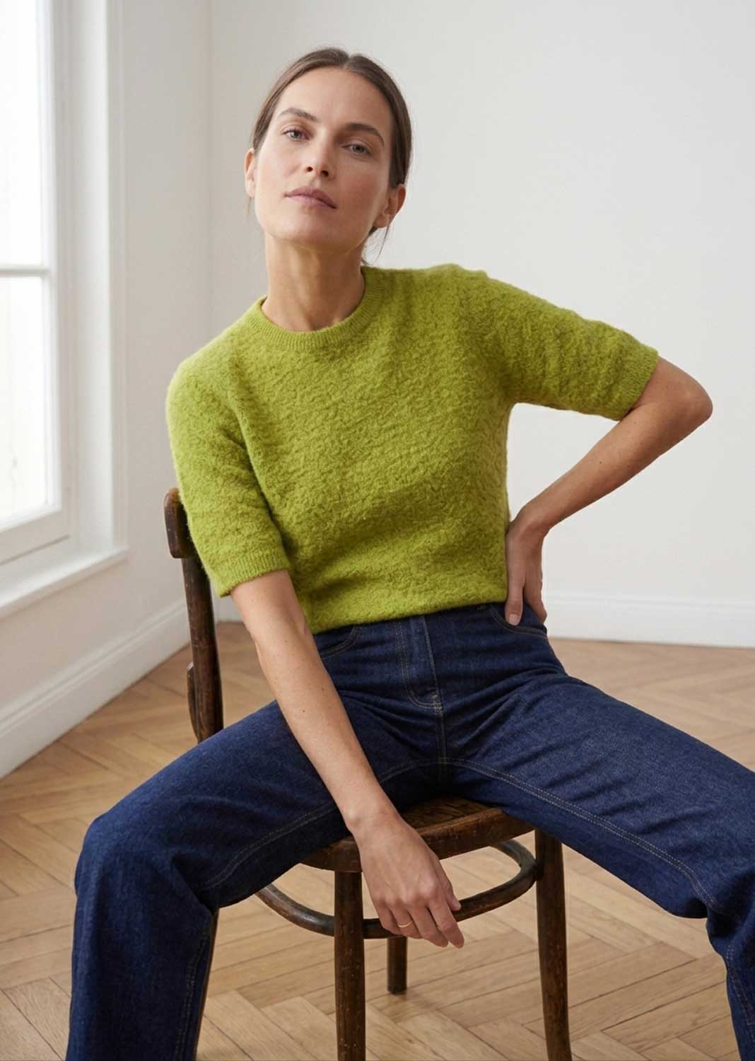 A woman sits on a wooden chair in a bright room, wearing a textured lime green short-sleeve knit top and dark blue jeans.