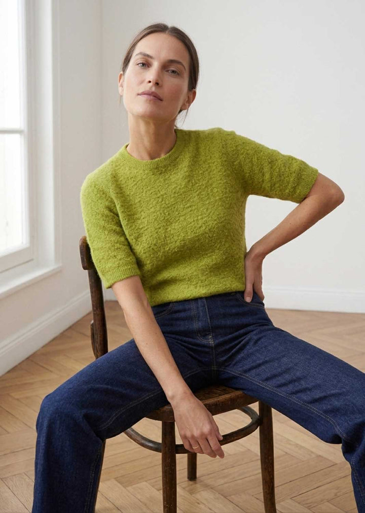 A woman sits on a wooden chair in a bright room, wearing a textured lime green short-sleeve knit top and dark blue jeans.