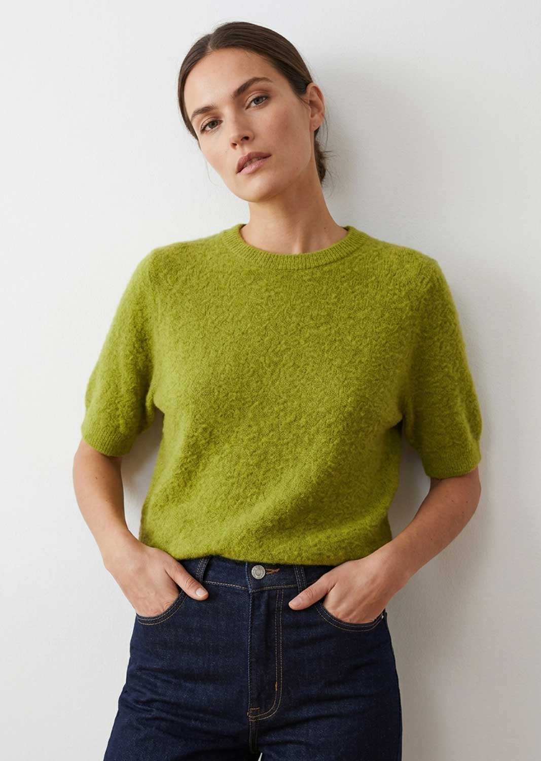 A woman wearing a short-sleeve, fuzzy lime green knit sweater and dark blue jeans, standing against a plain light gray wall.