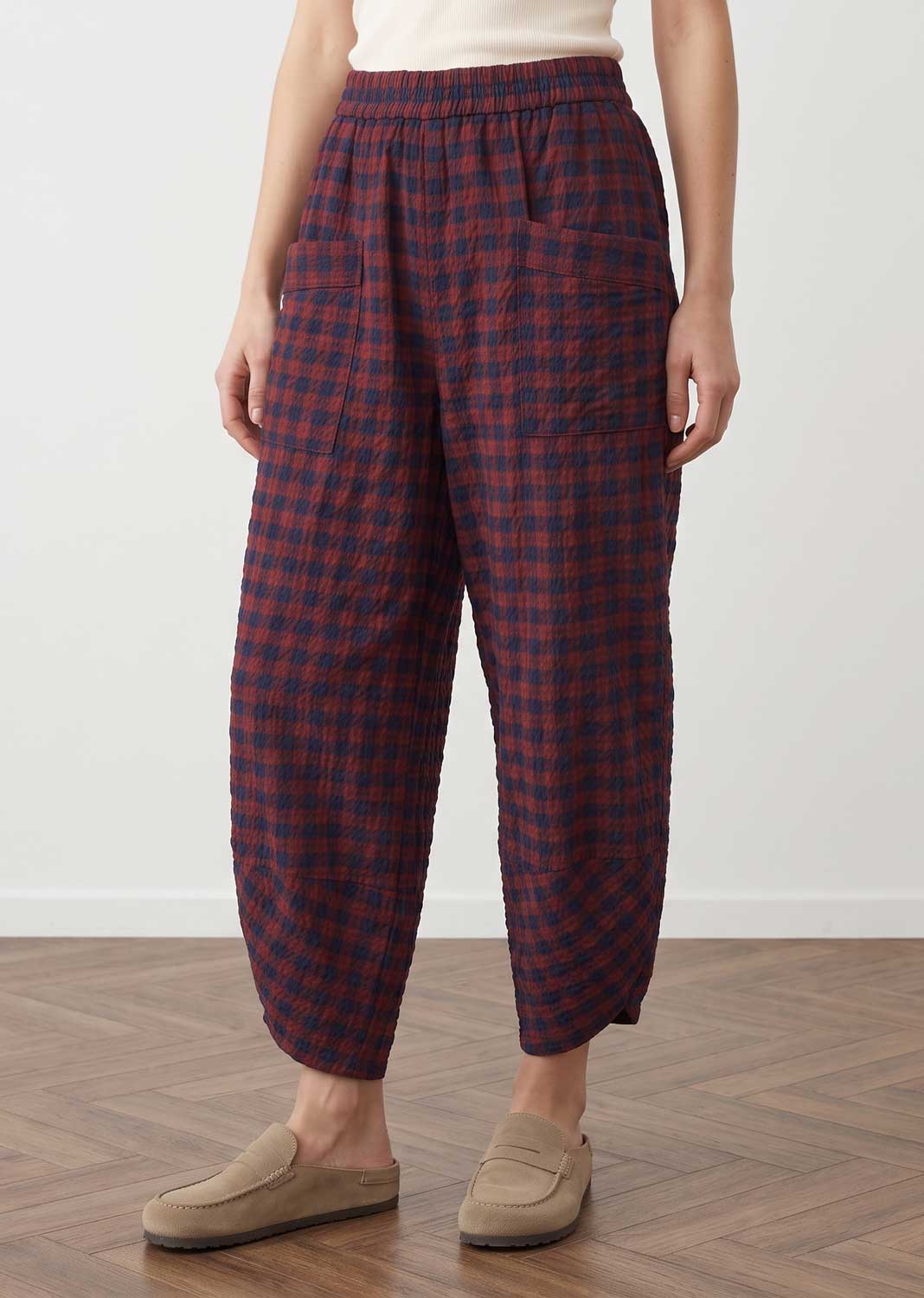 A person wearing loose, quilted ankle-length pants with a red and navy plaid pattern, paired with beige slip-on loafers, standing on a wood floor against a plain light wall.