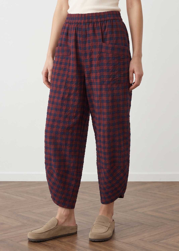 A person wearing loose, quilted ankle-length pants with a red and navy plaid pattern, paired with beige slip-on loafers, standing on a wood floor against a plain light wall.
