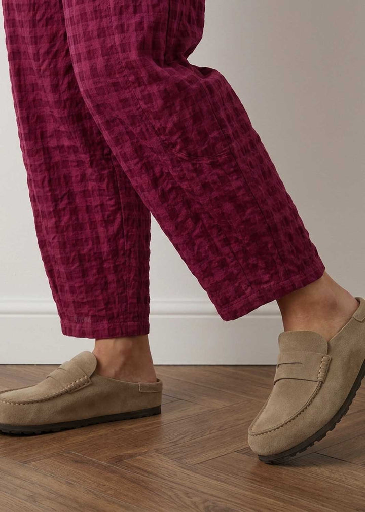 A person wearing textured magenta-purple cropped trousers and beige suede moccasin-style loafers, standing on a wood floor against a light neutral wall.