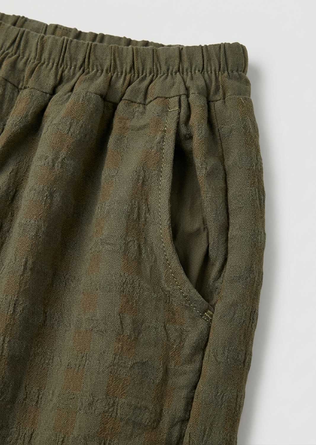 Close-up of olive green textured trousers with an elastic waistband and side pocket, shot against a light gray background.