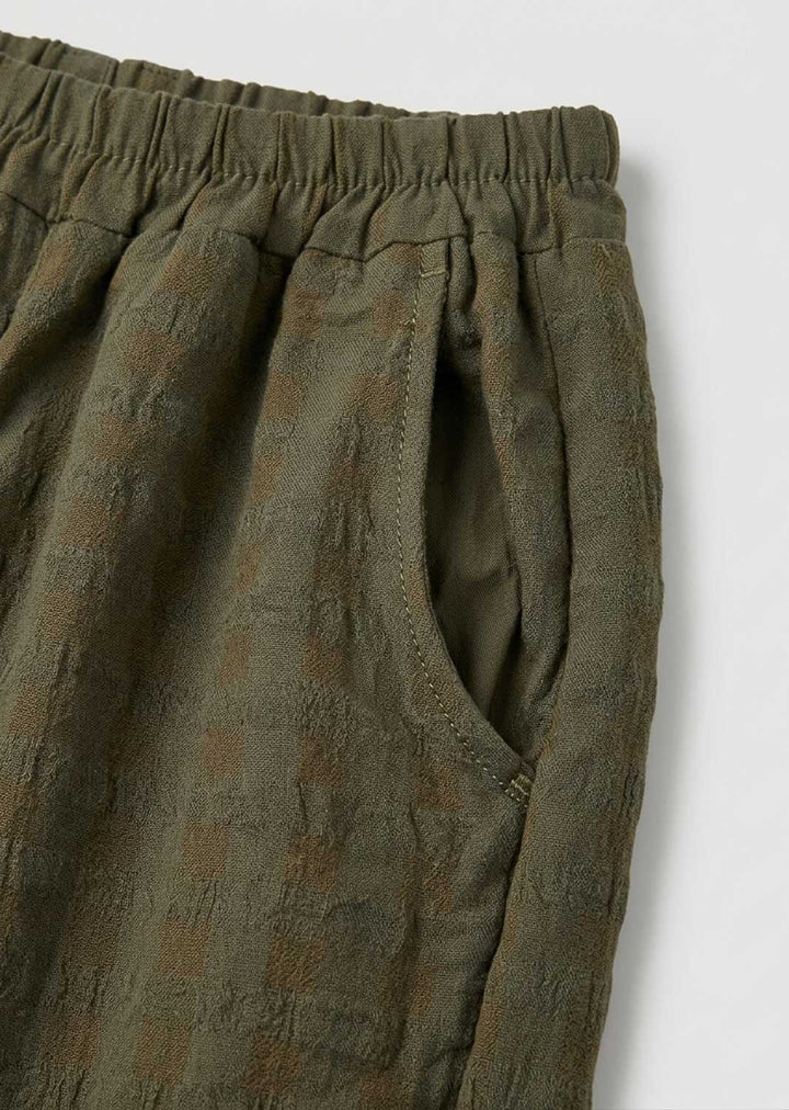 Close-up of olive green textured trousers with an elastic waistband and side pocket, shot against a light gray background.