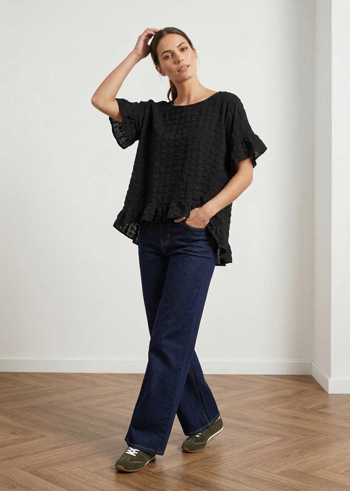 A woman modeling a black textured short-sleeve blouse with ruffled sleeves and hem, paired with dark blue jeans and olive green sneakers, standing in a minimalist room with a light gray wall and wooden parquet floor.