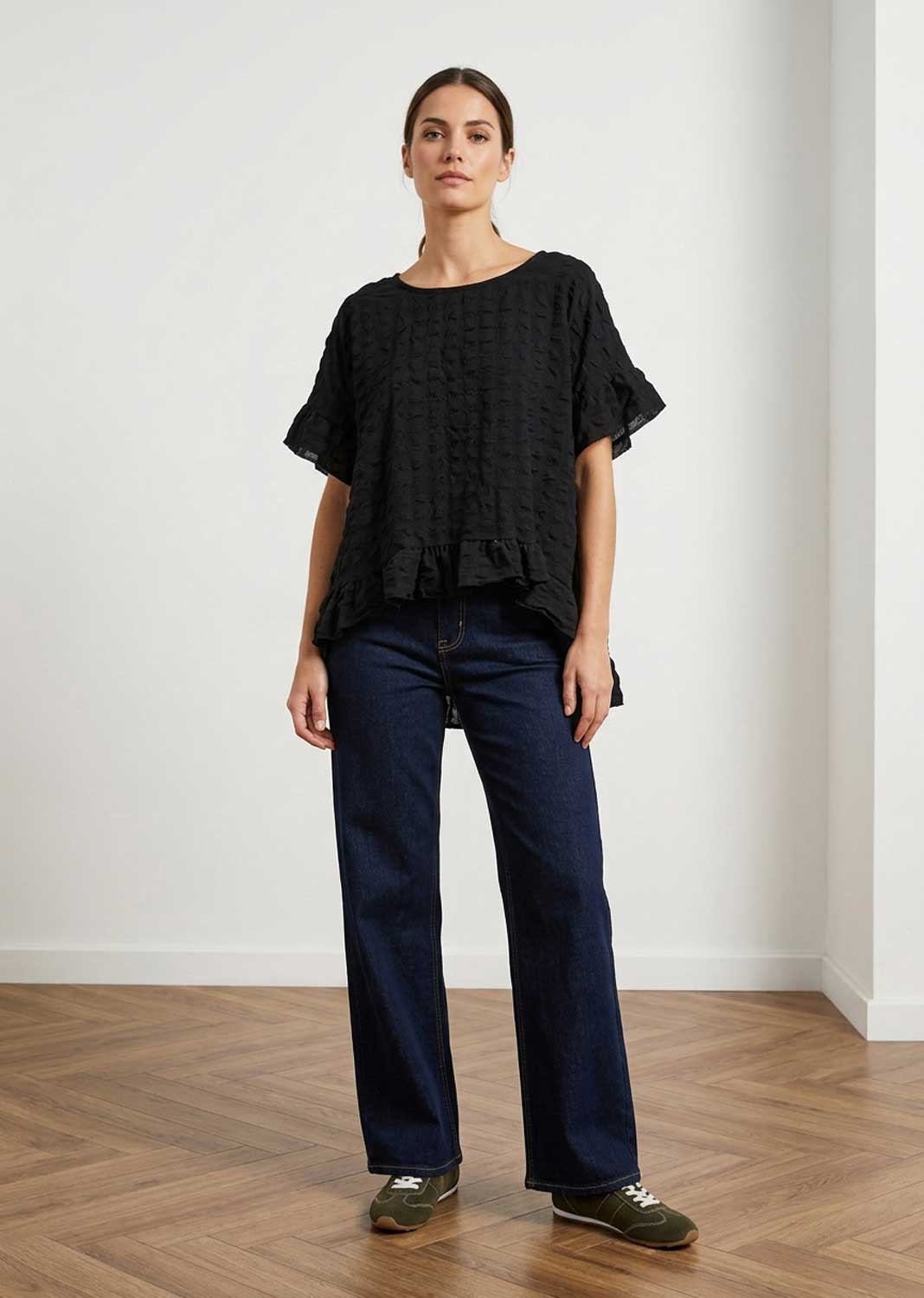 A woman stands in a minimalist room with a light gray wall and wood floor, wearing a black textured seersuker short-sleeve blouse with ruffled details, dark blue wide-leg jeans, and olive green sneakers.
