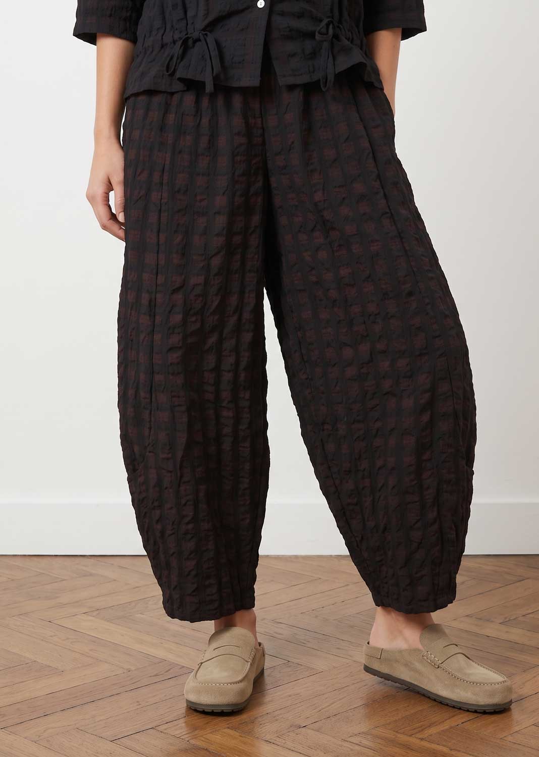 A person wearing voluminous black textured harem-style pants with a subtle tonal pattern, paired with a black top and beige slip-on shoes, standing on a wooden floor against a plain white wall.