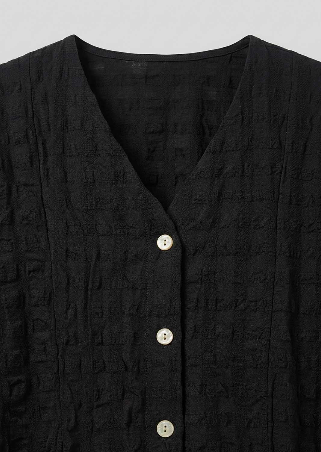 Close-up of a black textured button-front top with a V-neck and light-colored buttons, shown against a plain light gray background.