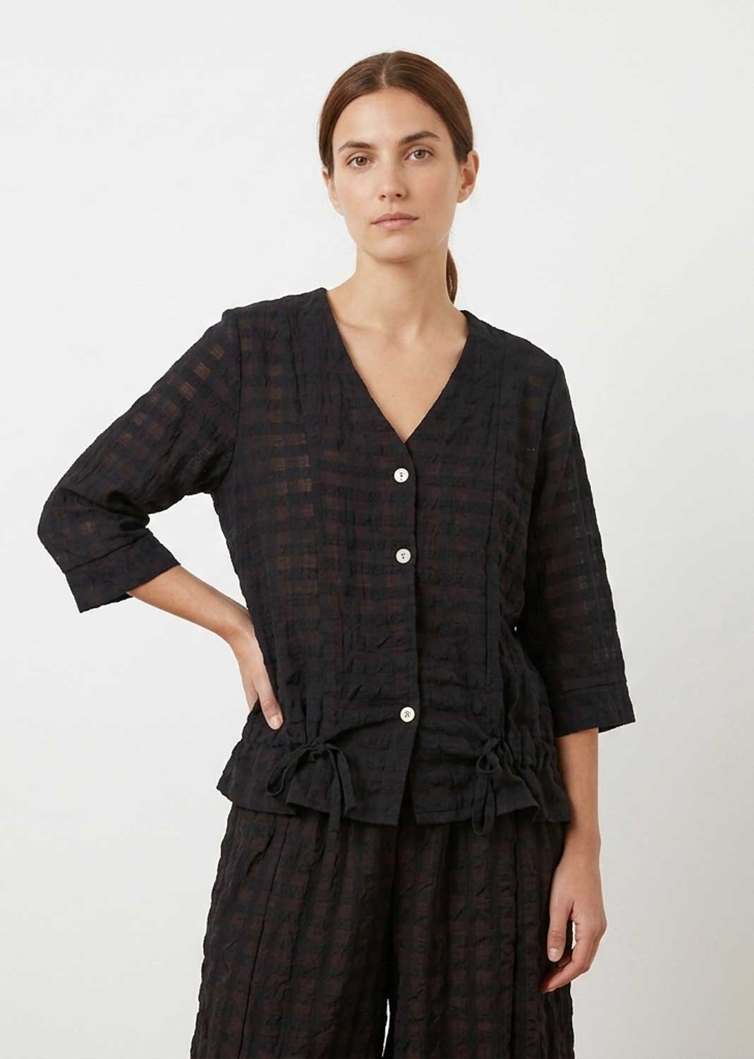 A woman modeling a black and dark brown checkered two-piece set consisting of a cropped button-front top with three-quarter sleeves and matching wide-leg trousers, shot against a plain light gray background.