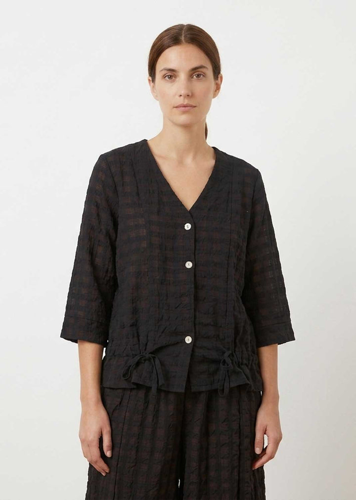 A woman modeling a black textured, patterned three-quarter sleeve top with a V-neck and front buttons, paired with matching dark trousers, against a plain light gray background.