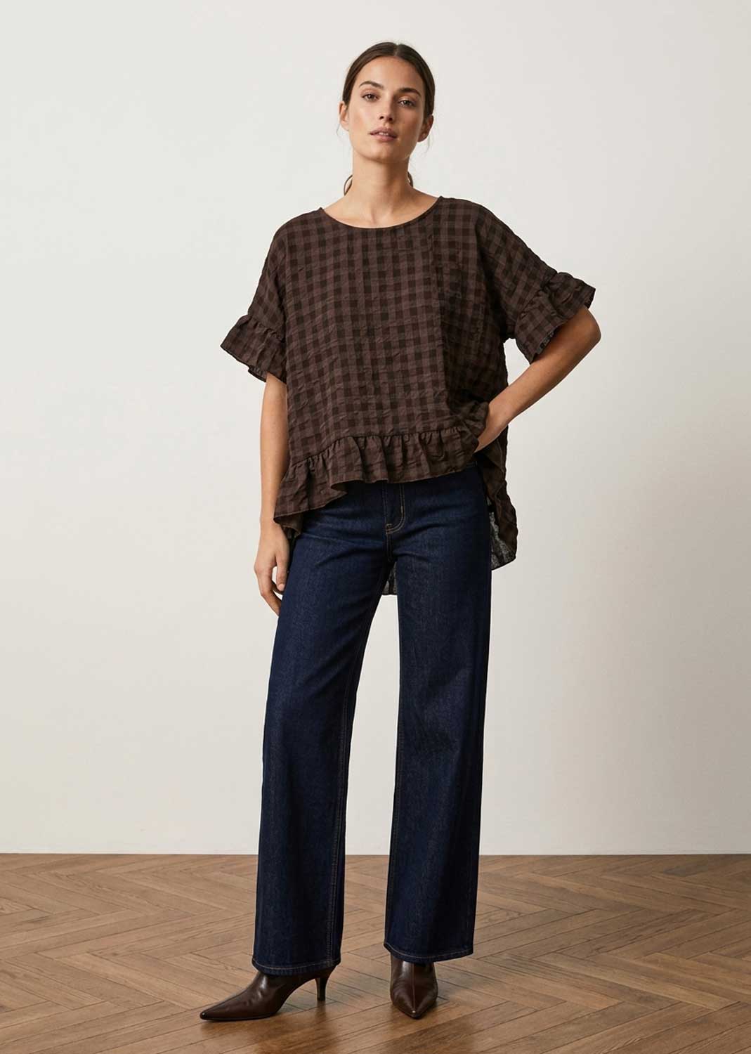 A woman modeling a dark brown  checkered short-sleeve blouse with ruffled sleeves and hem, paired with dark blue wide-leg jeans and brown pointed-toe heels, standing against a neutral off-white wall and wood herringbone floor.