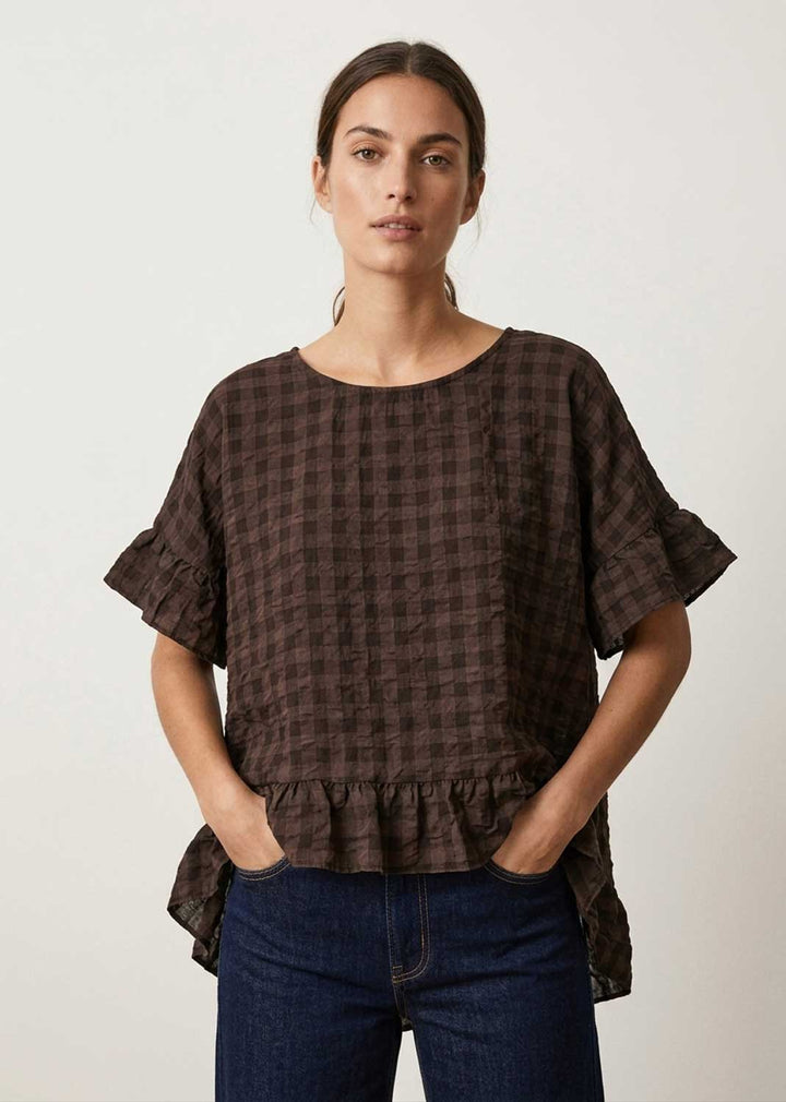 A woman modeling a dark brown checkered short-sleeve blouse with ruffled sleeves and a ruffled hem, paired with dark blue jeans, against a plain light gray background.