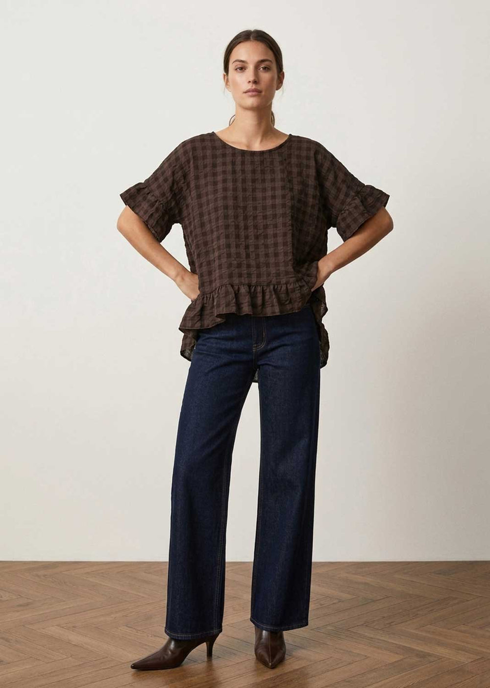 A woman stands in a studio wearing a dark brown and black checkered short-sleeve blouse with a ruffled hem, paired with dark blue wide-leg jeans and brown pointed-toe heels.
