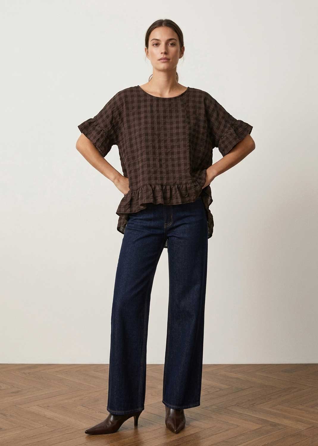 A woman stands in a studio wearing a dark brown and black checkered short-sleeve blouse with a ruffled hem, paired with dark blue wide-leg jeans and brown pointed-toe heels.