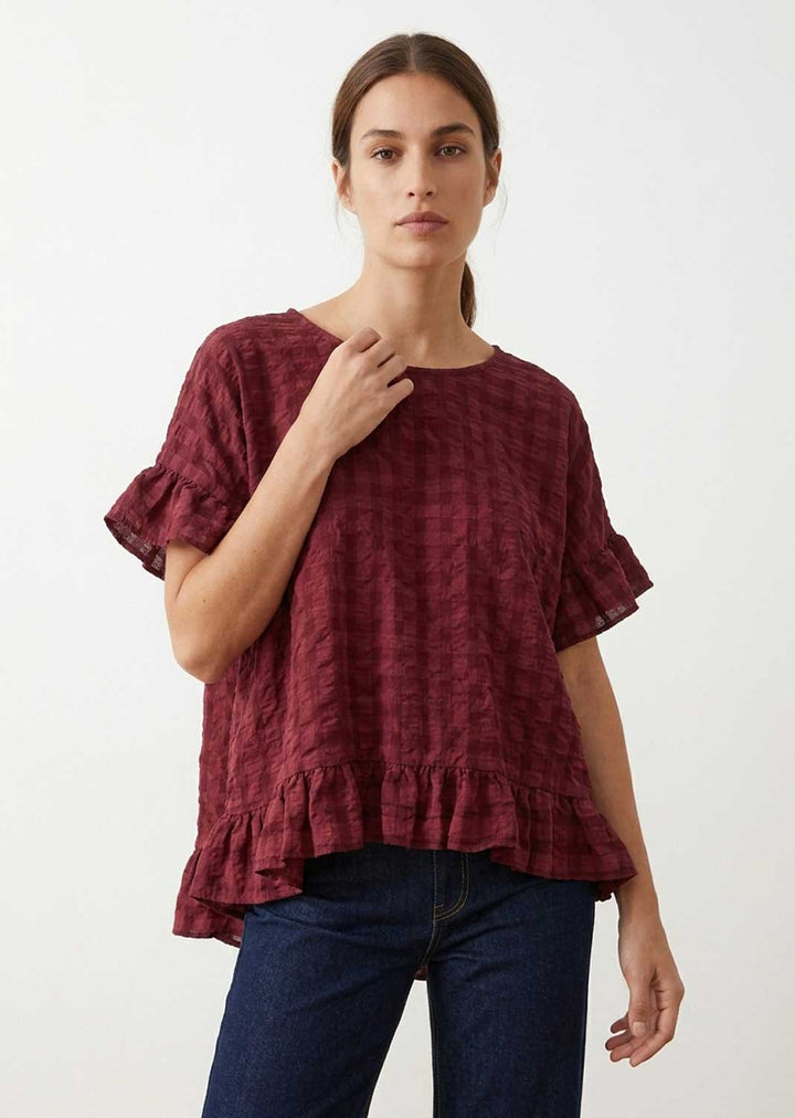 A woman wearing a textured, short-sleeved top in a deep burgundy red with ruffled details, paired with dark blue jeans, against a plain white background.
