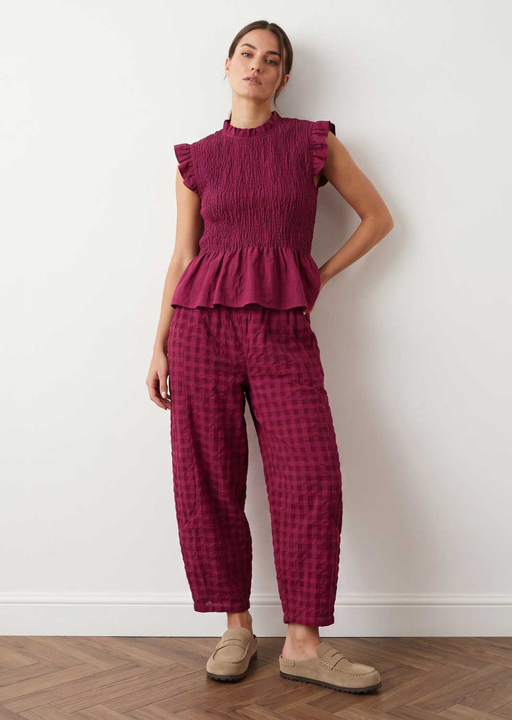 A woman stands against a white wall wearing a matching two-piece outfit with a smocked peplum top and wide-leg plaid pants in deep magenta, paired with beige slip-on shoes.