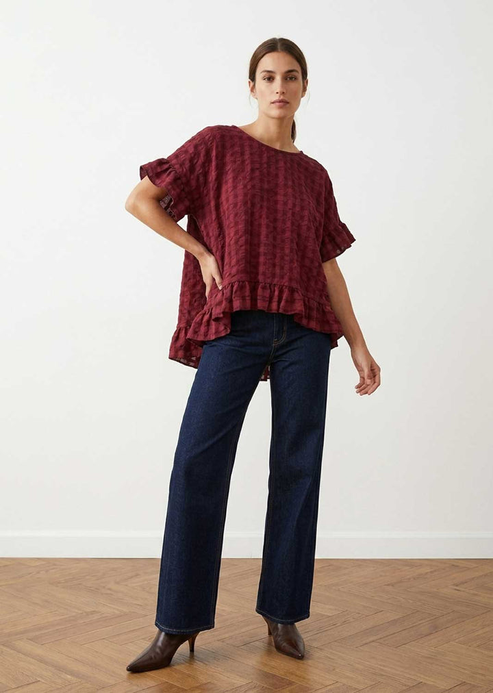A woman modeling a loose-fitting, short-sleeved burgundy top with a subtle checkered pattern and ruffled details, paired with dark blue jeans and brown pointed-toe heels, standing in a minimalist room with a white wall and wood floor.