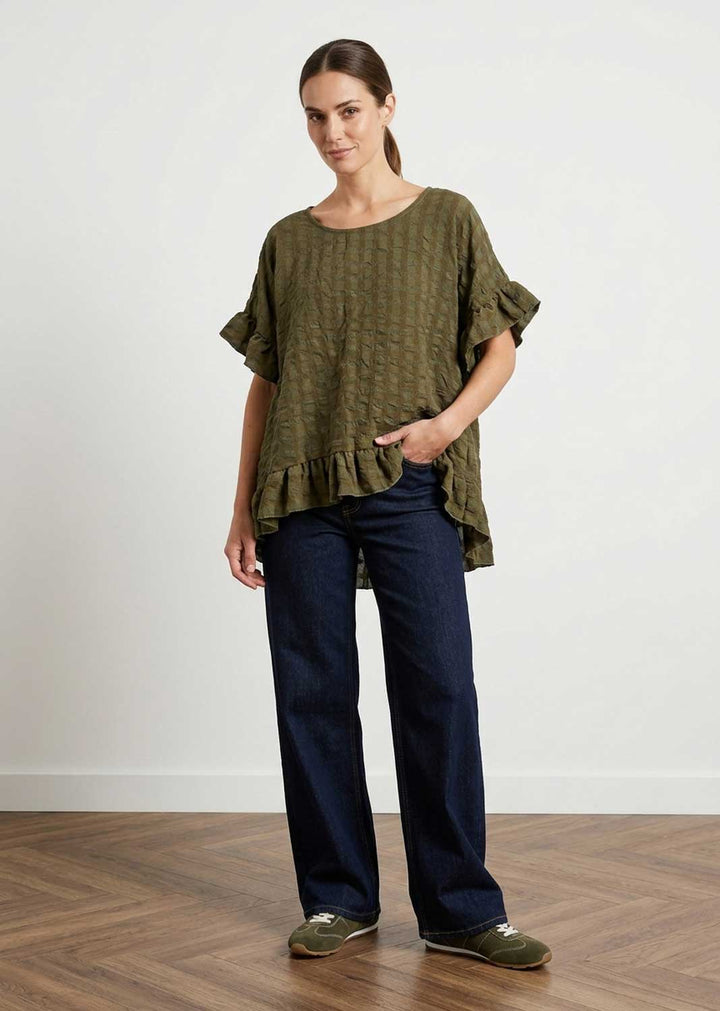 A woman modeling a loose seersucker olive green short-sleeve top with ruffled sleeves and hem, paired with dark blue jeans and olive green slip-on shoes, standing on a wooden floor against a white wall.