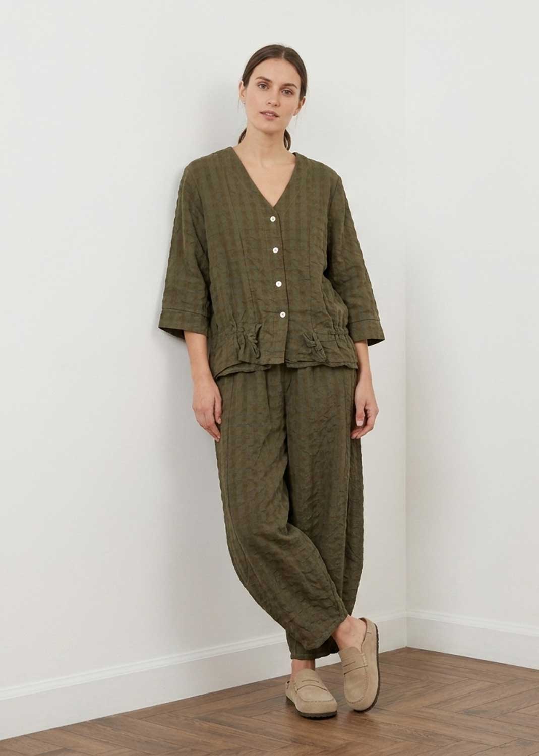 A woman modeling a matching olive green two-piece linen set consisting of a cropped button-front top and wide-leg trousers, paired with beige slip-on shoes, standing against a white wall.