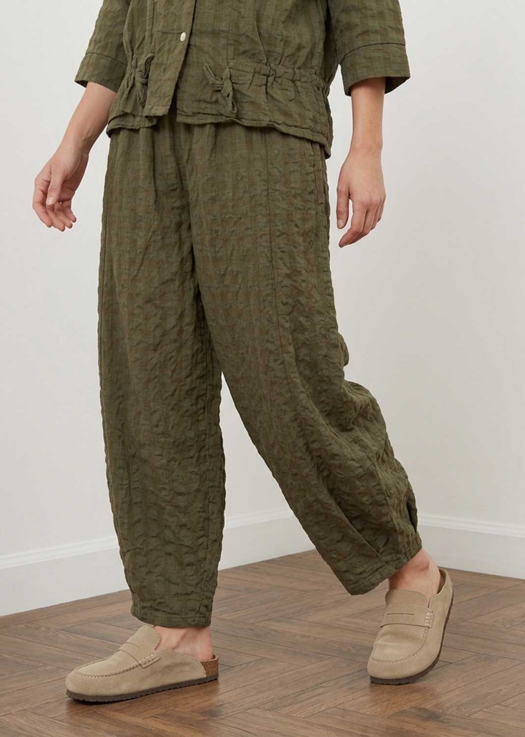 A person wearing a matching olive green textured two-piece outfit with a cropped button-front top and loose wide-leg pants, paired with beige slip-on loafers, standing on a wood floor against a plain light wall.