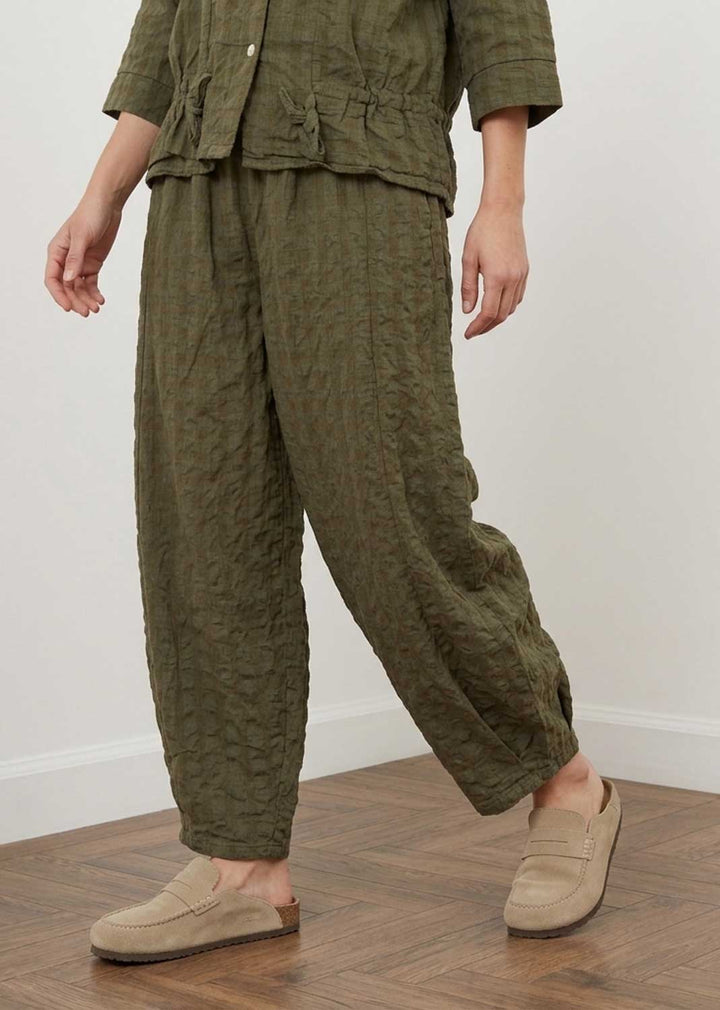 A person wearing a matching olive green textured two-piece outfit with a cropped button-front top and loose wide-leg pants, paired with beige slip-on loafers, standing on a wood floor against a plain light wall.
