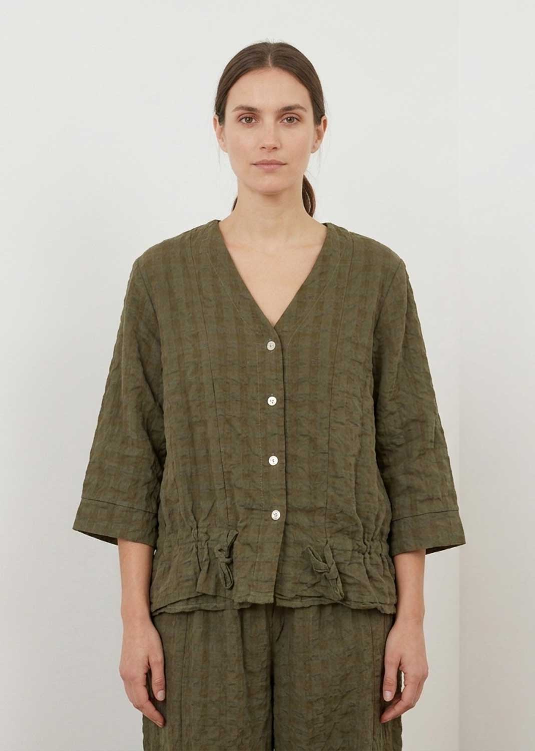 A woman modeling a textured olive green two-piece linen set consisting of a cropped button-front top and matching trousers, shot against a plain off-white background.