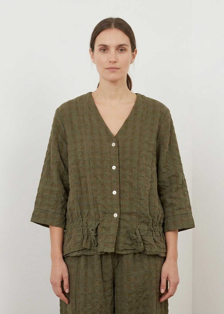 A woman modeling a textured olive green two-piece linen set consisting of a cropped button-front top and matching trousers, shot against a plain off-white background.