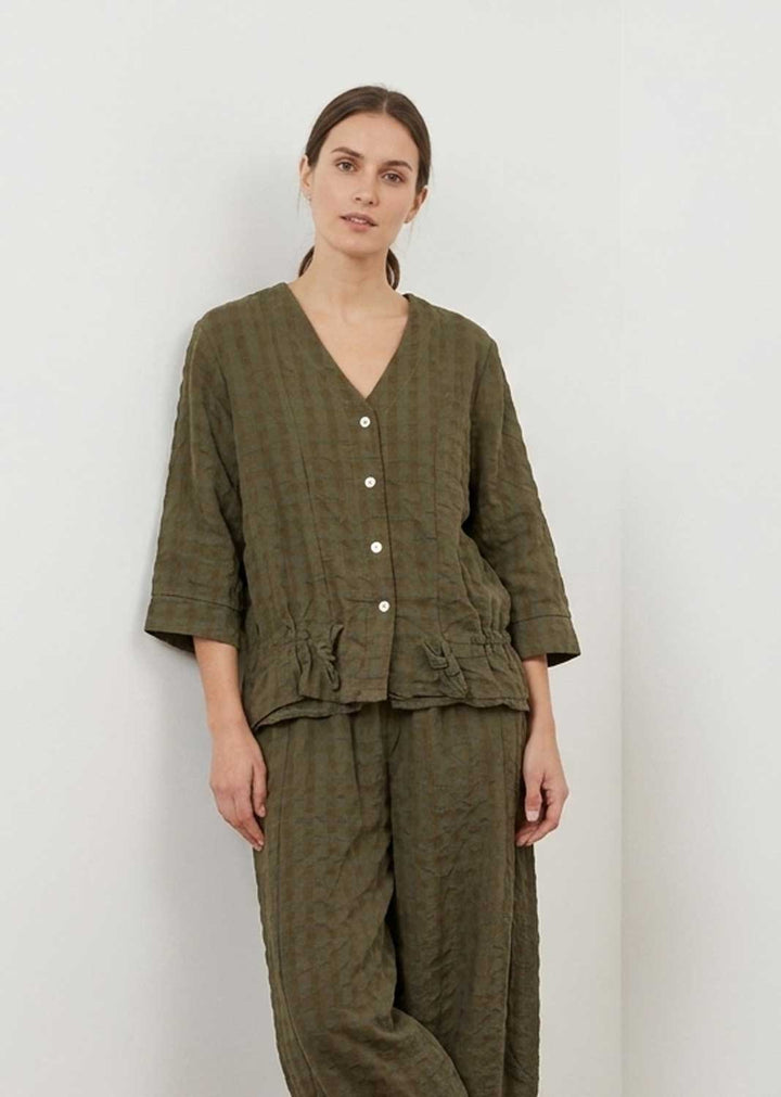 A woman modeling a matching olive green textured two-piece lounge set consisting of a short-sleeve button-front top and wide-leg pants, set against a plain light gray wall.