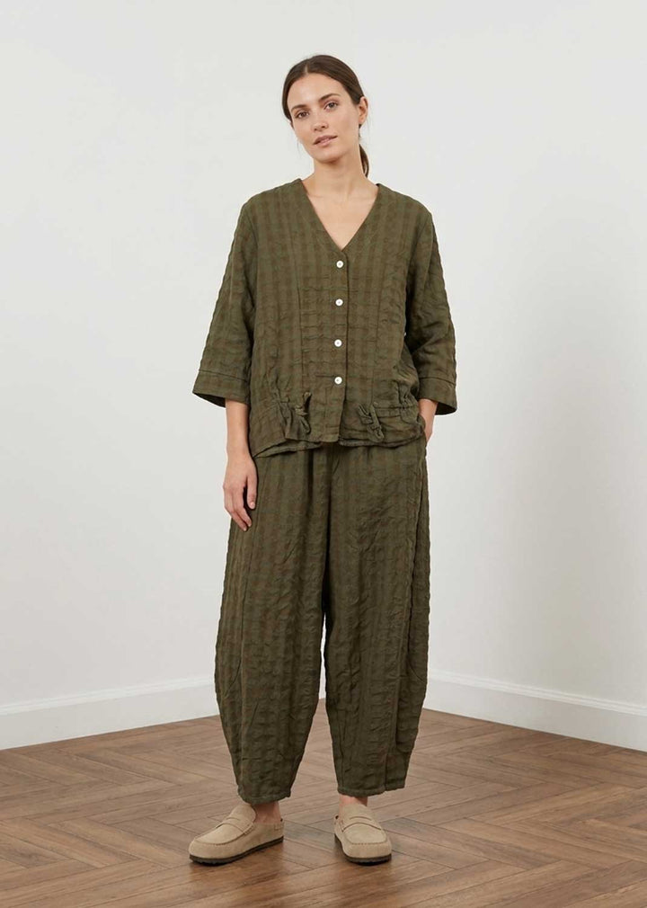 A woman modeling a matching olive green textured two-piece lounge set consisting of a button-front long-sleeve top and wide-leg pants, paired with beige slip-on shoes, in a minimalist room with a white wall and wood floor.