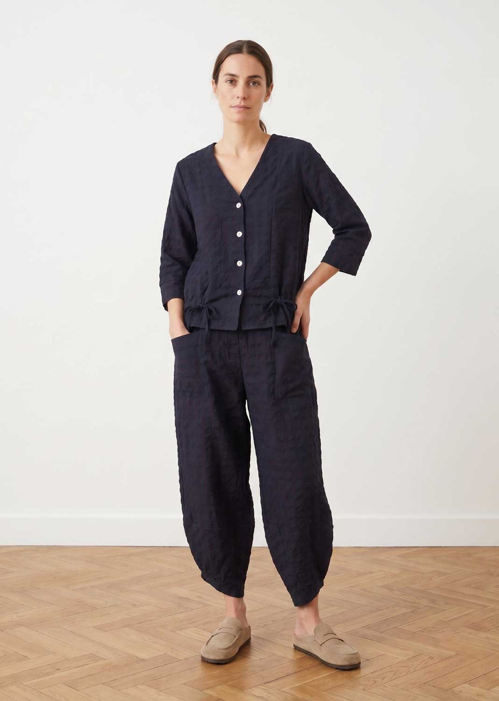 A woman stands in a minimalist room with a light wall and wood parquet floor, wearing a dark navy blue matching set consisting of a button-front long-sleeve top and wide-leg cropped pants, paired with beige slip-on shoes.