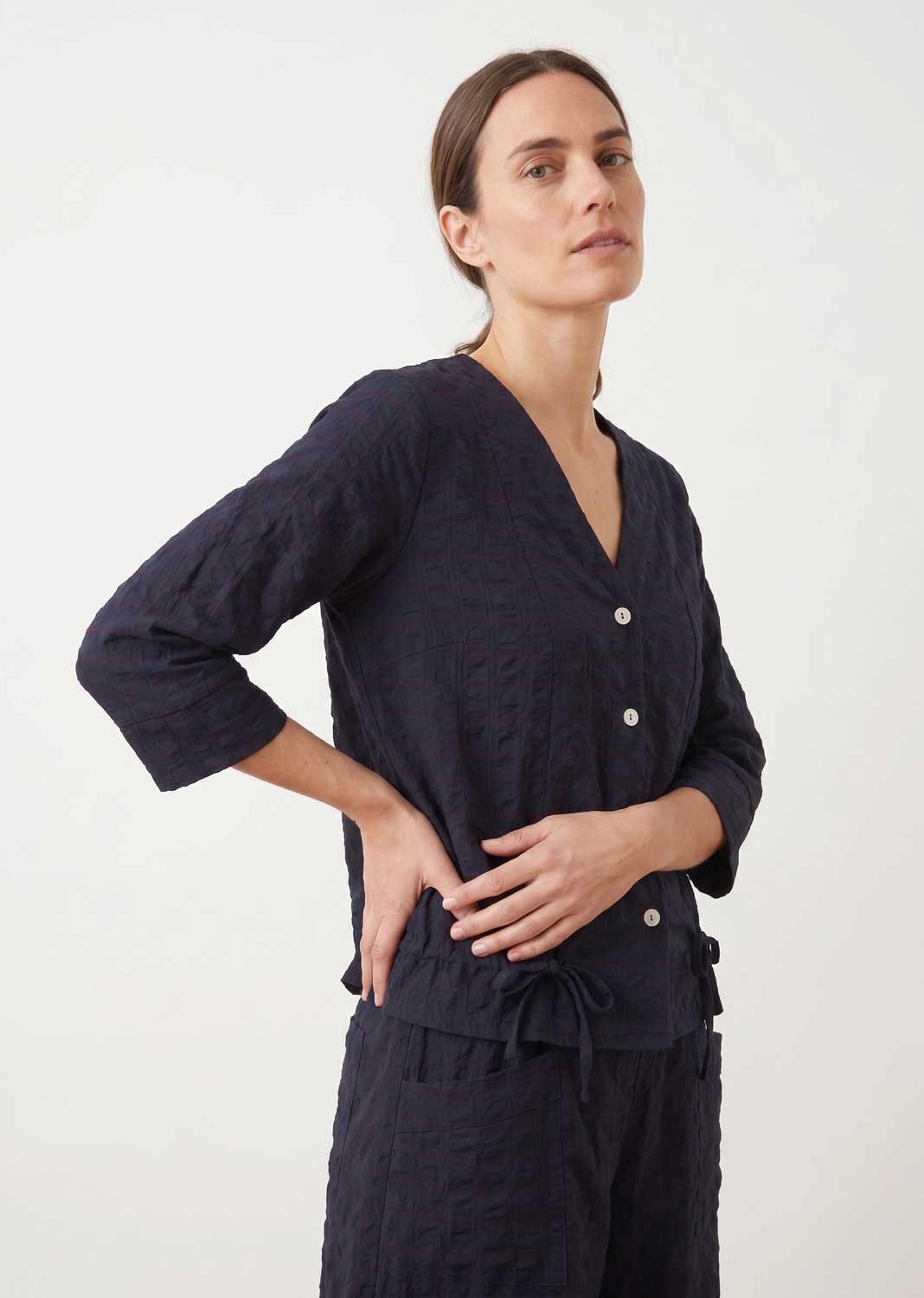 A woman modeling a dark navy textured two-piece set consisting of a button-front long-sleeve top and matching drawstring pants, photographed against a clean off-white studio background.
