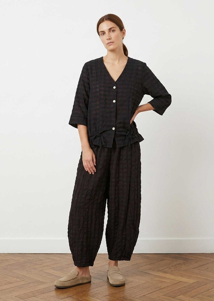 A woman modeling a black and dark brown checkered two-piece set consisting of a cropped button-front top with three-quarter sleeves and matching wide-leg pants, paired with beige slip-on shoes against a white wall and wood floor.