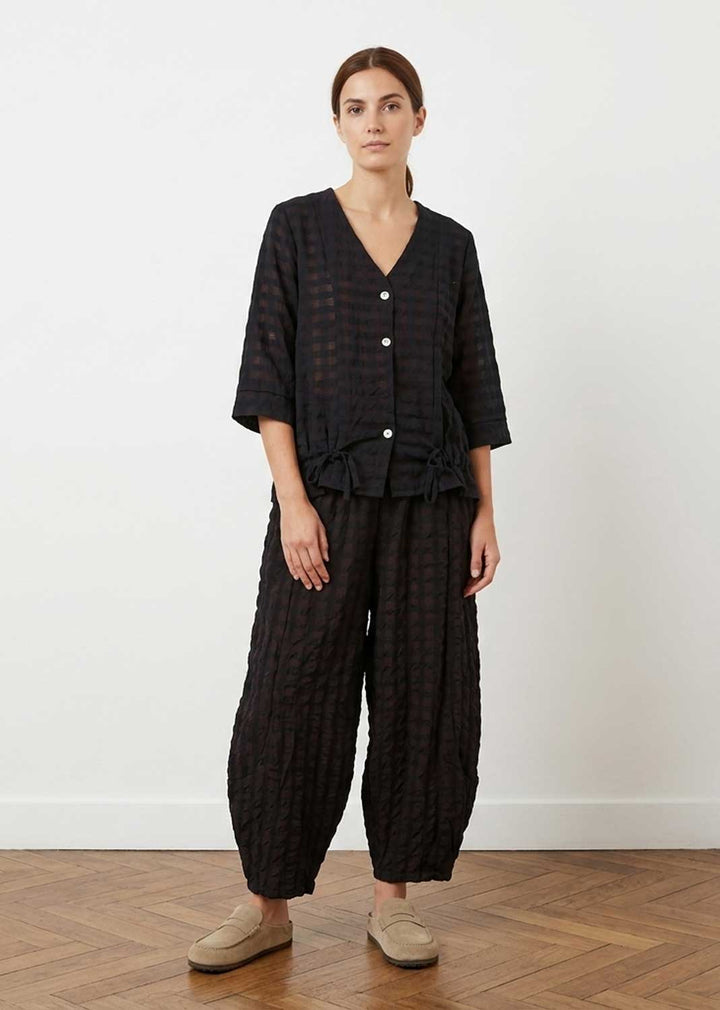 A woman stands in a studio wearing a loose black textured two-piece outfit with a button-front top and matching wide-leg pants, paired with beige slip-on shoes against a plain light wall and wood floor.