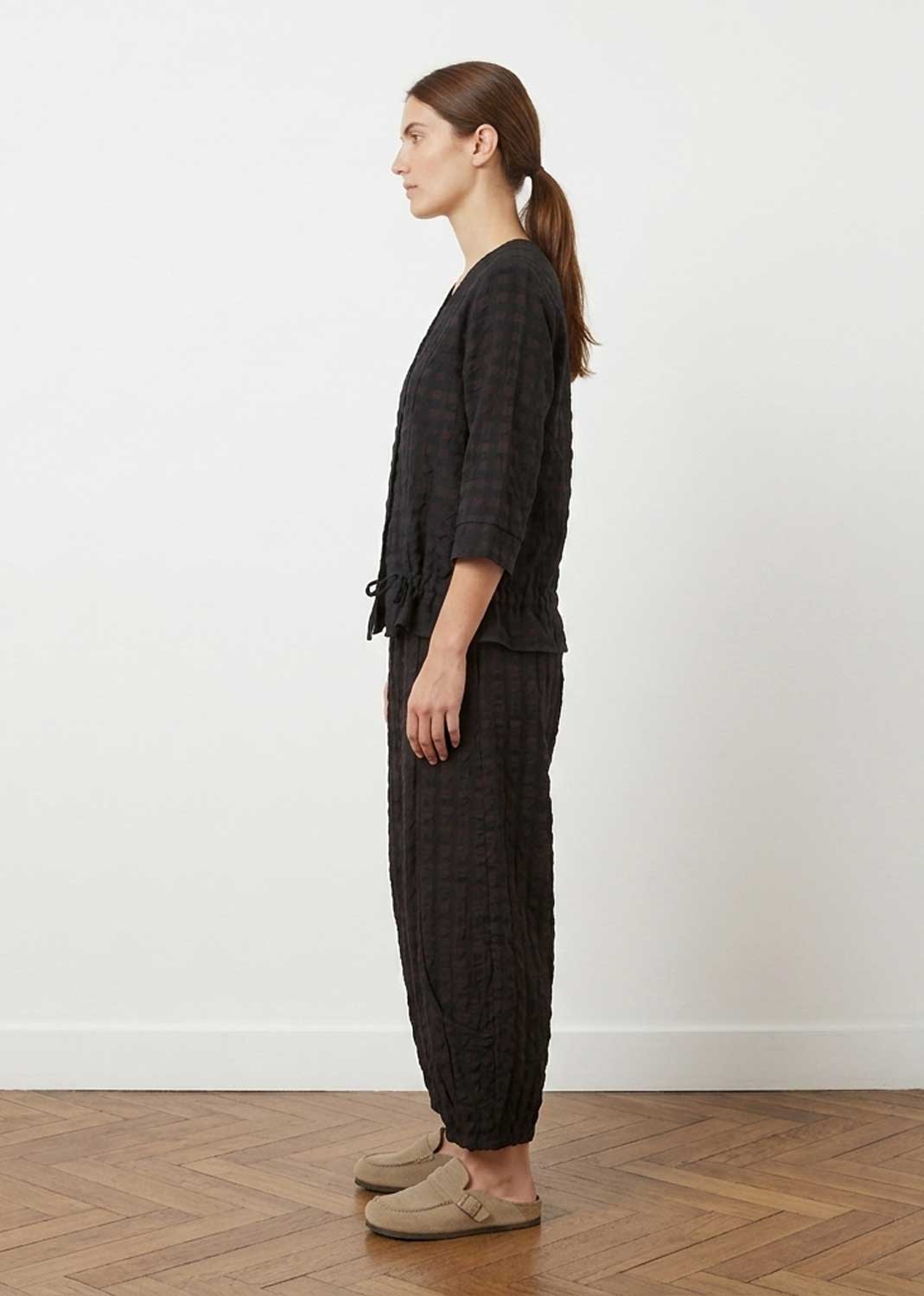 A woman stands in profile wearing a matching dark charcoal plaid lounge set consisting of a cropped long-sleeve top and wide-leg pants, paired with beige slip-on shoes against a white wall and wood floor.