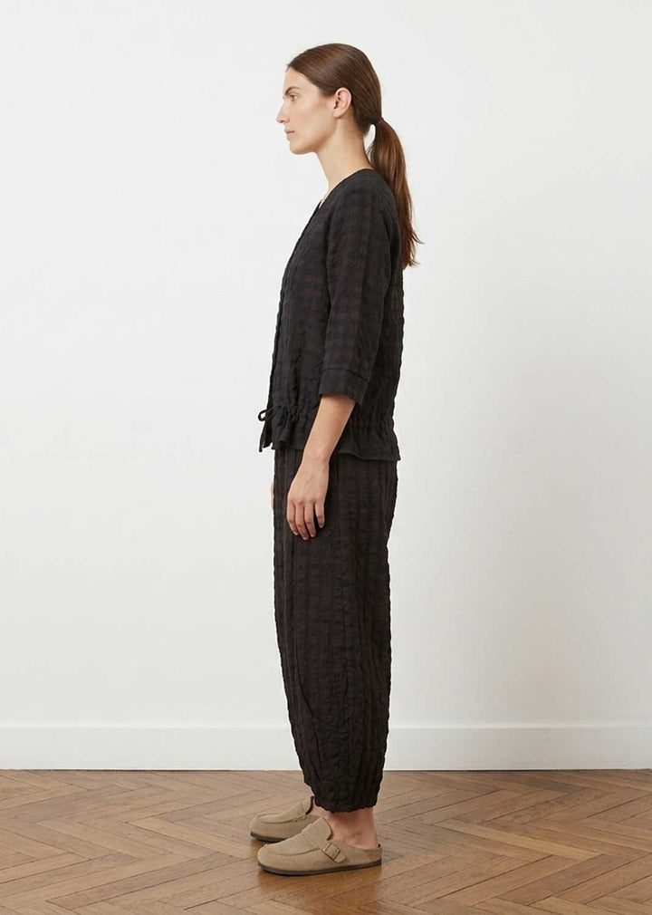 A woman stands in profile wearing a matching dark charcoal plaid lounge set consisting of a cropped long-sleeve top and wide-leg pants, paired with beige slip-on shoes against a white wall and wood floor.