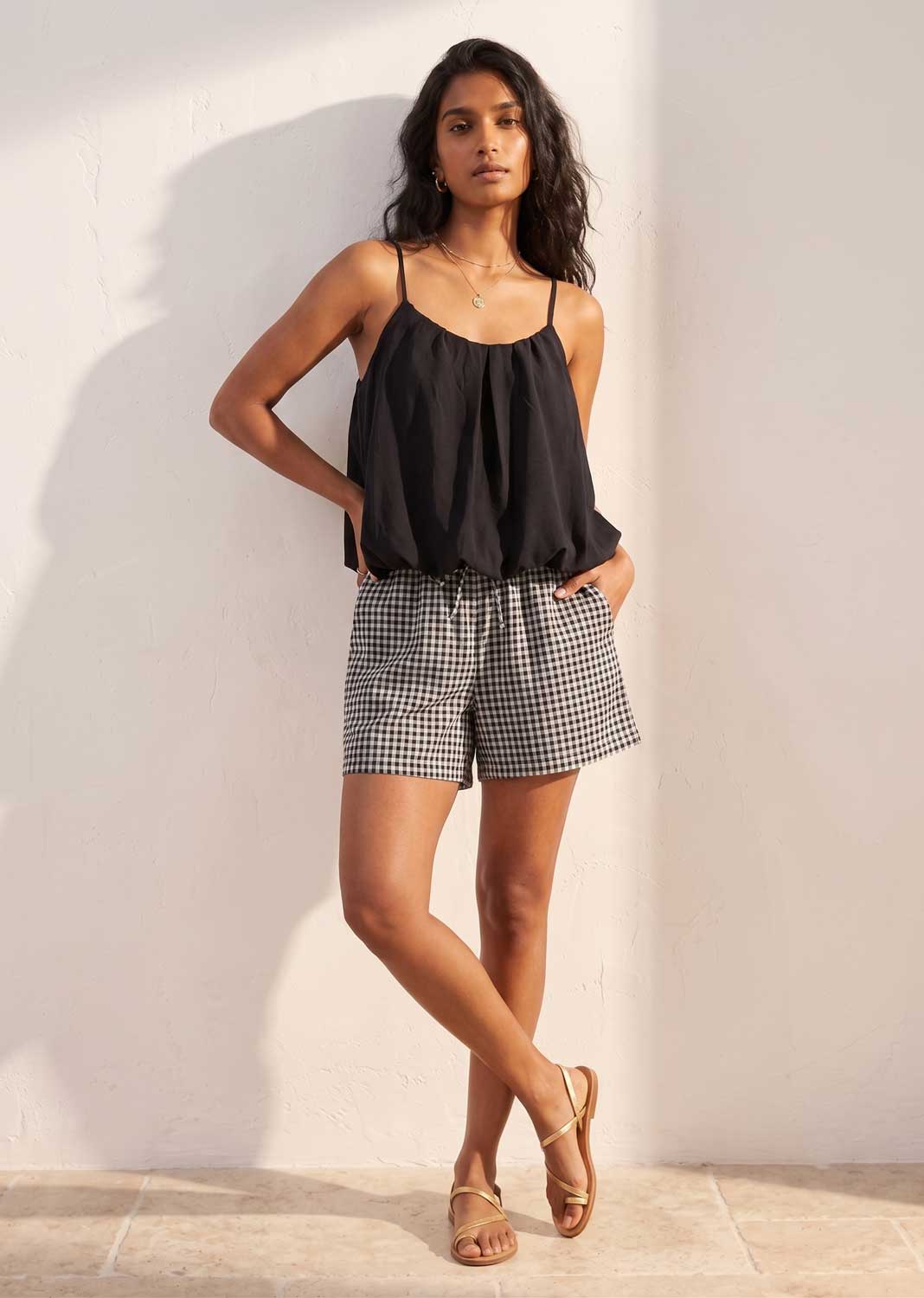 Woman wearing a black top and gingham checkered shorts against a white wall.