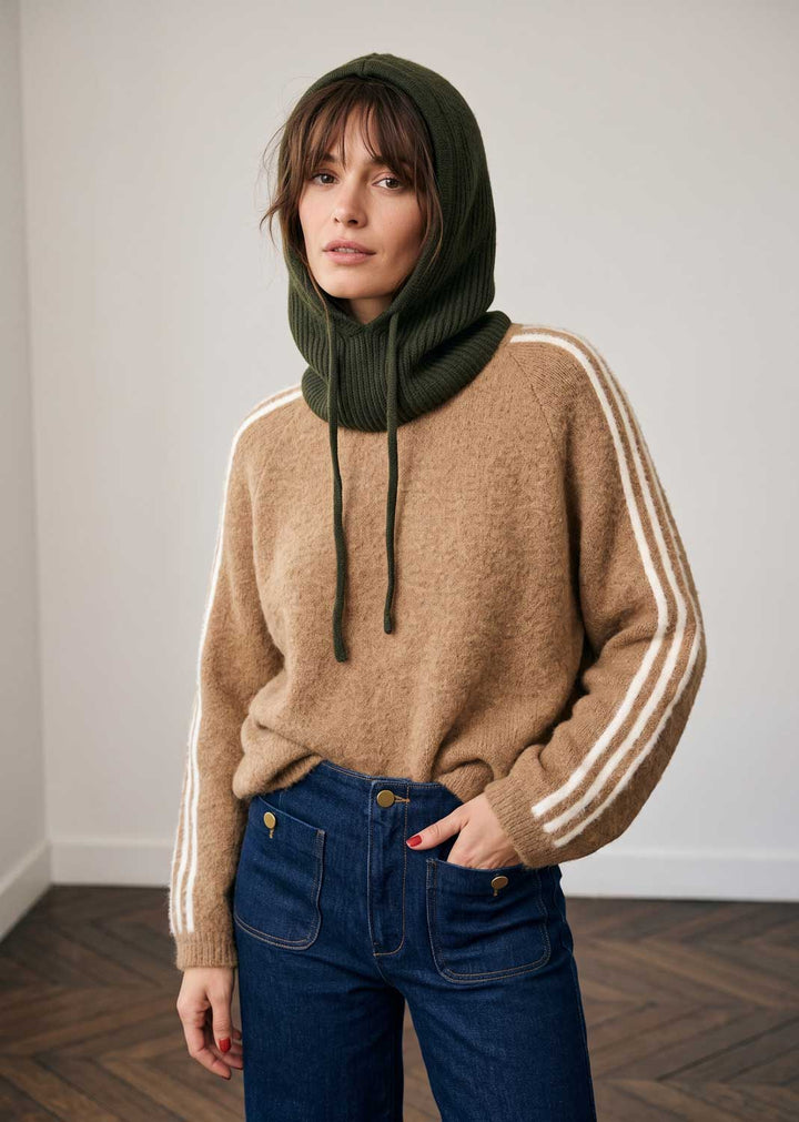 A woman wearing a beige knit sweater with white stripes on the sleeves, a dark olive green ribbed cowl neck hood, and blue jeans, standing in a minimalist room with a light gray wall and wood floor.