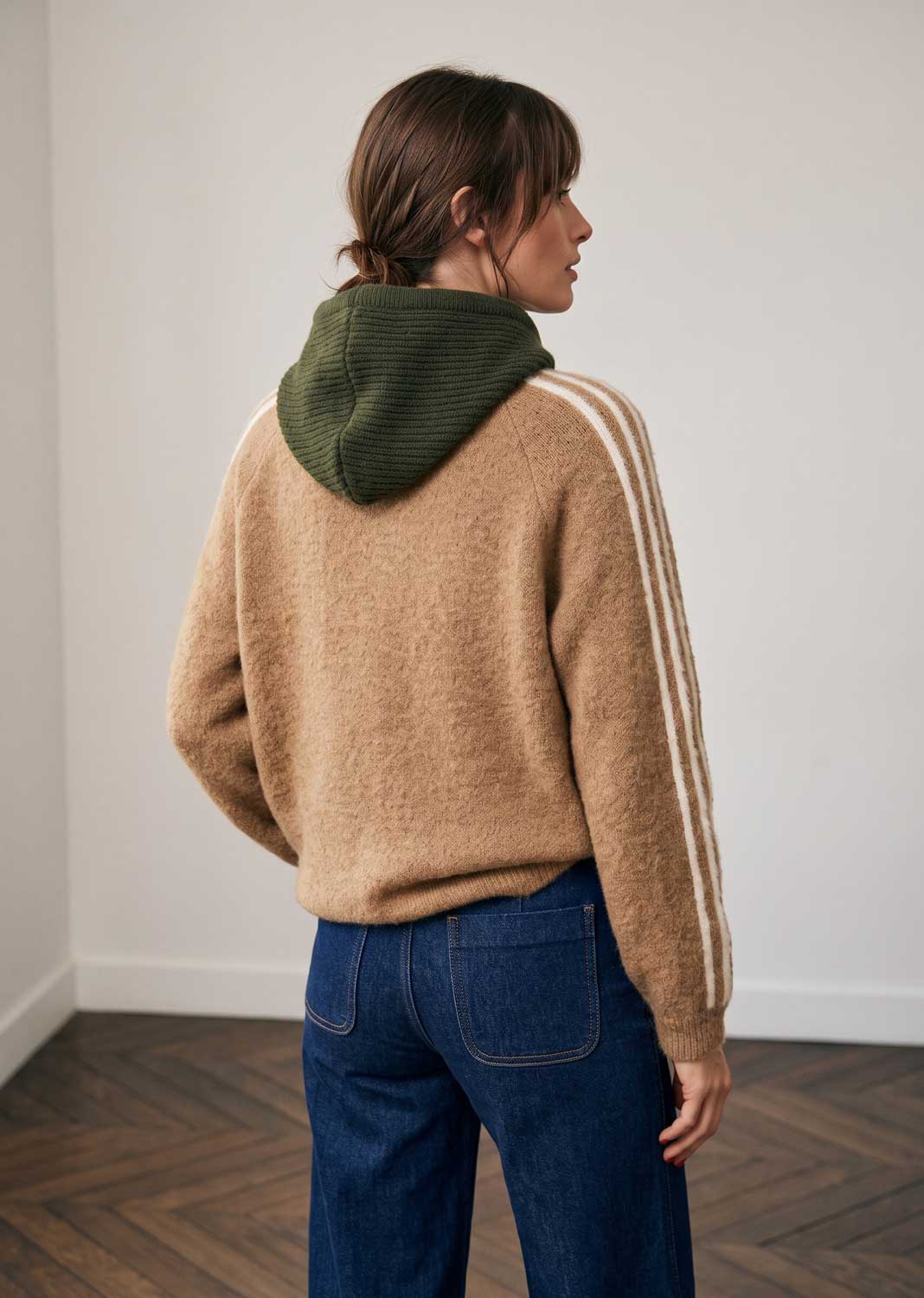 A woman shown from the back wearing a beige fuzzy knit sweater with an olive green ribbed hood and white stripes down the sleeves, paired with blue jeans in a minimalist room with a wood floor.
