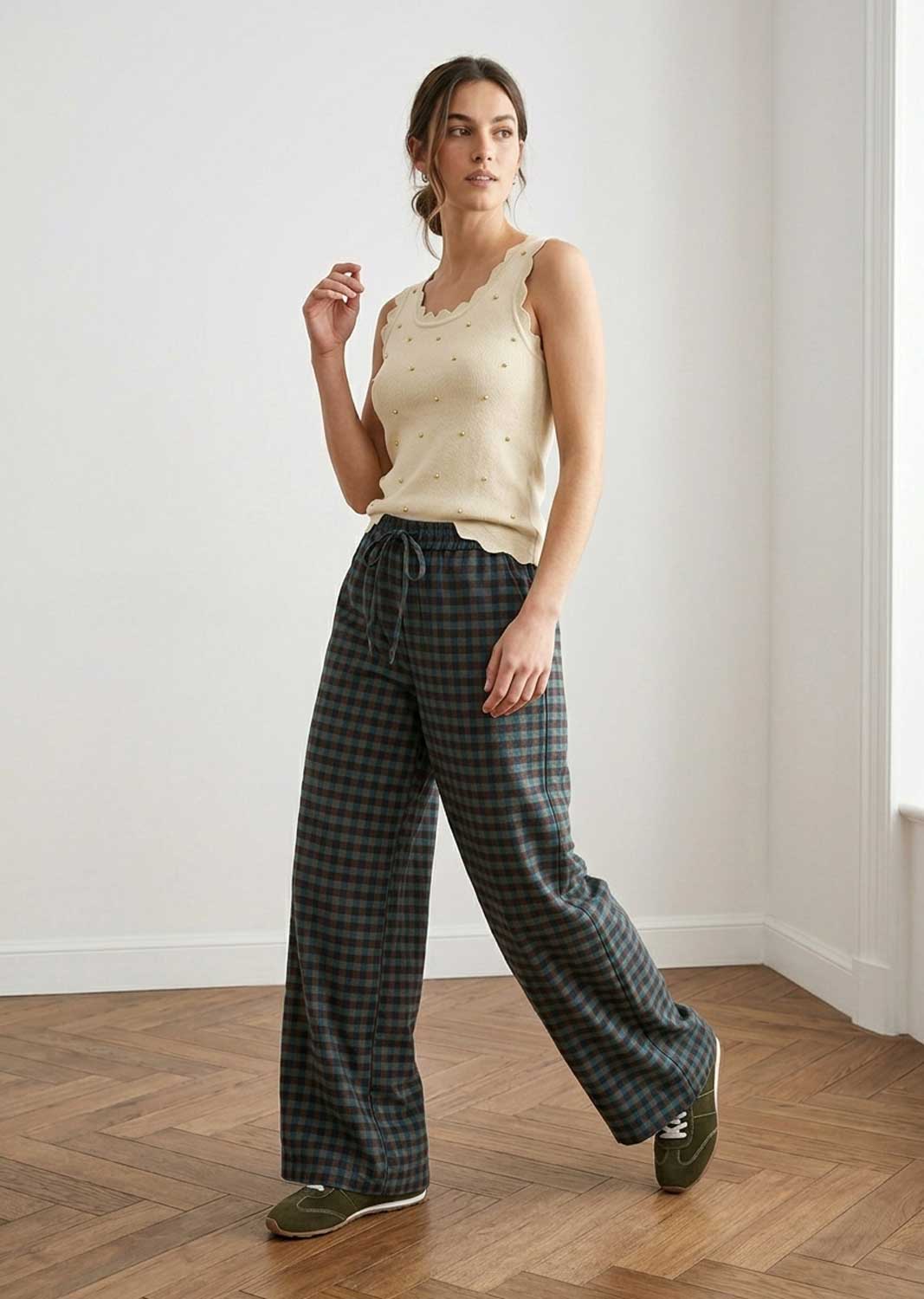 A woman modeling a cream sleeveless knit top with delicate bead details and dark green plaid wide-leg trousers, standing in a bright room with a wood herringbone floor.