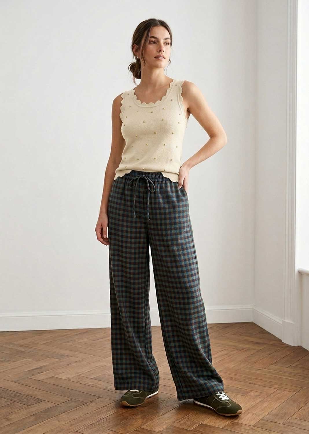 A woman stands in a bright room with a wood floor and white walls, wearing a cream sleeveless top with scalloped edges and small embroidered details, paired with dark green and black plaid wide-leg trousers and olive sneakers.