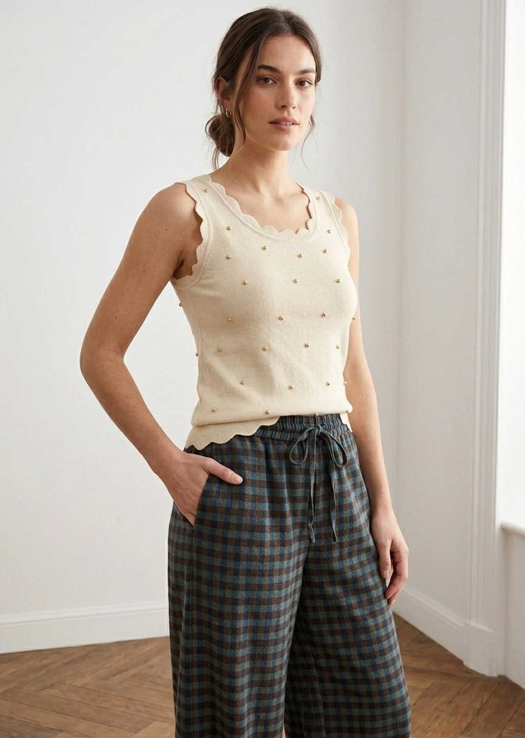 A woman modeling a cream sleeveless knit top with scalloped edges and small gold dot details, paired with dark green and gray plaid wide-leg trousers, standing in a bright room with white walls and wood flooring.